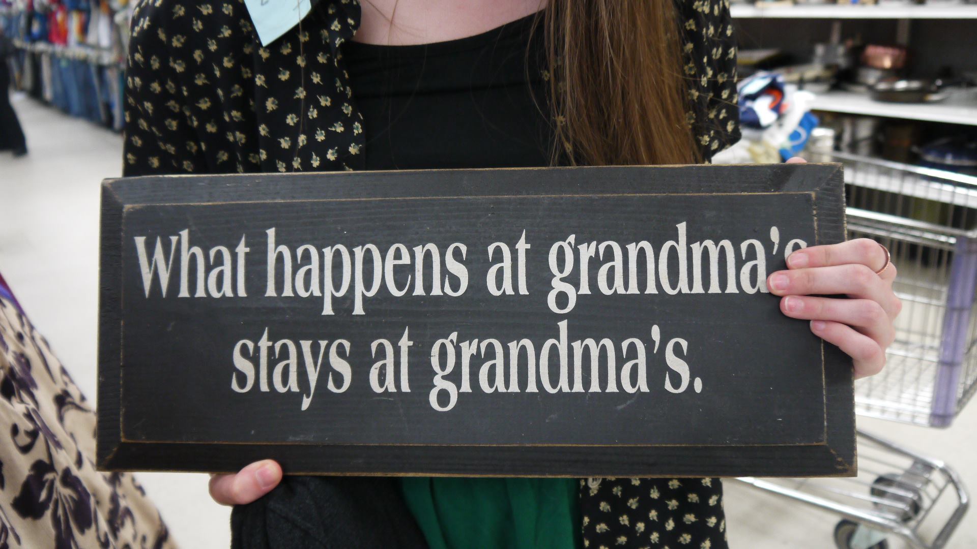 Individual holding wall art in thrift store that says - What happens at grandma's stays at grandma's.