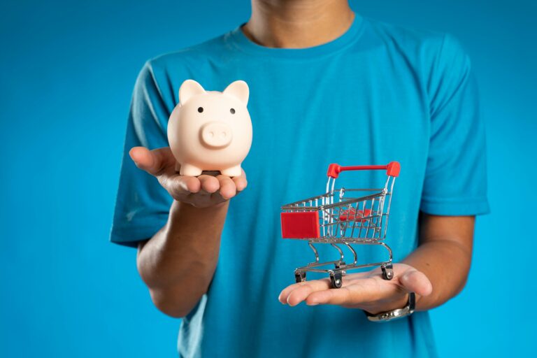 Individual holding a small piggy bank in one hand and a small shopping cart in the other
